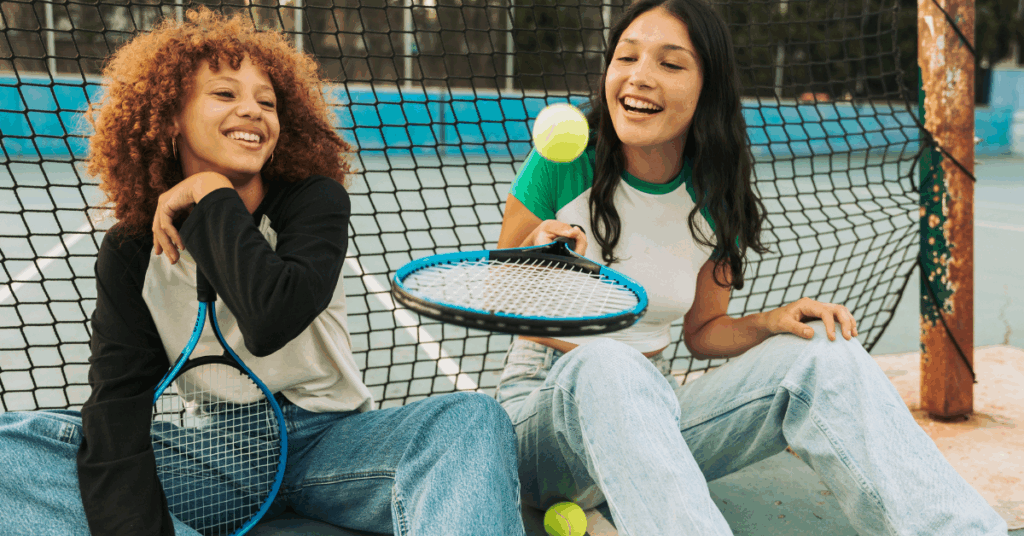 Two young girls in trendy outfits sitting in front of a tennis net bouncing the ball